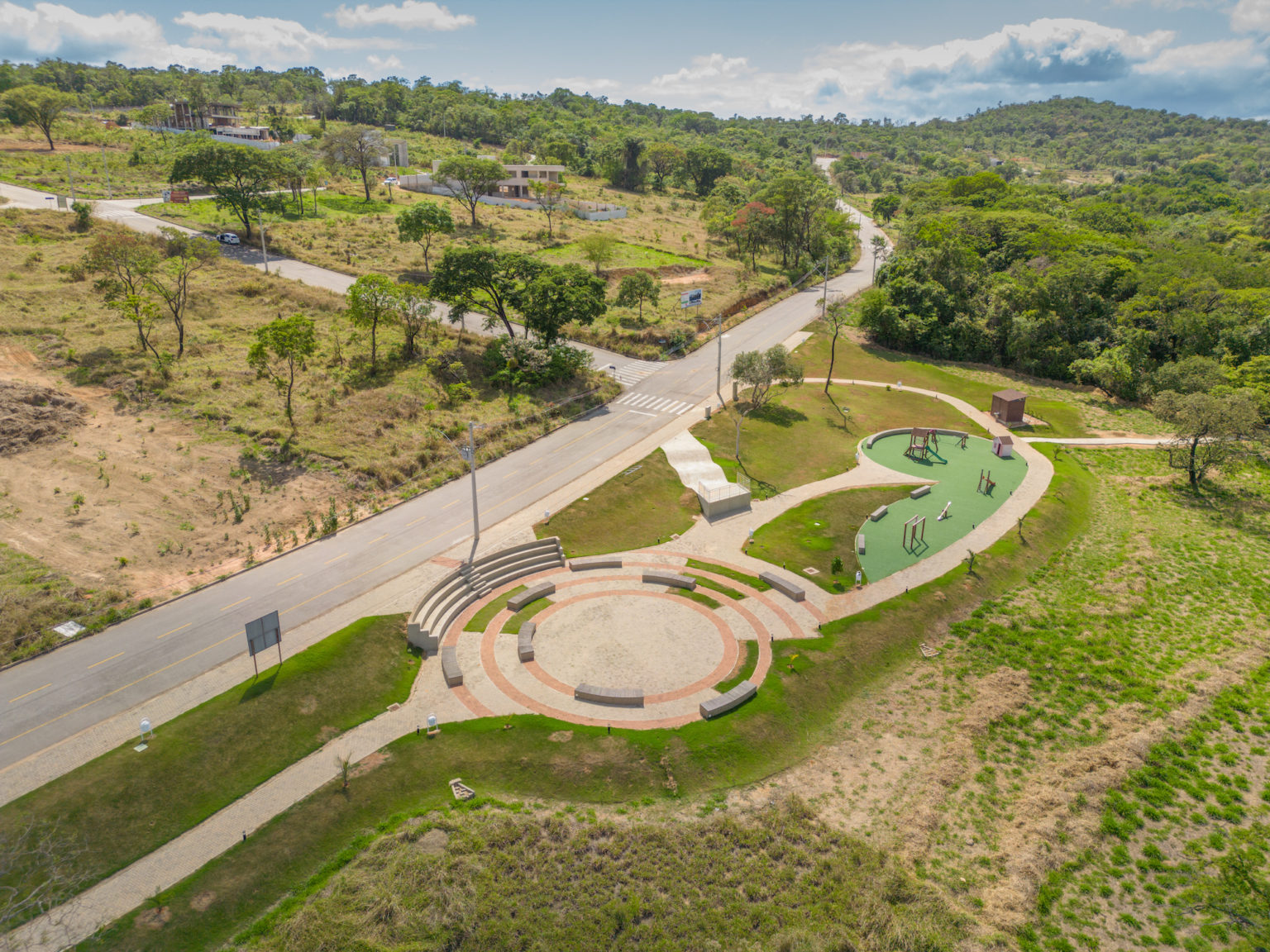 Foto da área de parquinho, com piso verde, pequeno mirante, área de anfiteatro, com arquibancada circular e mais alguns bancos rodeando o centro e pequeno quiosque de apoio