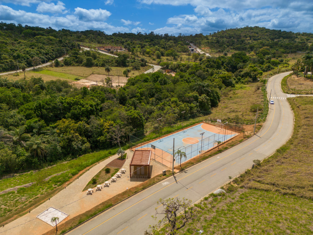 Foto de uma quadra de futebol e basquete em concreto com pintura laranja e azul gradeada. Ao lado quiosque de apoio com algumas mesas em área externa