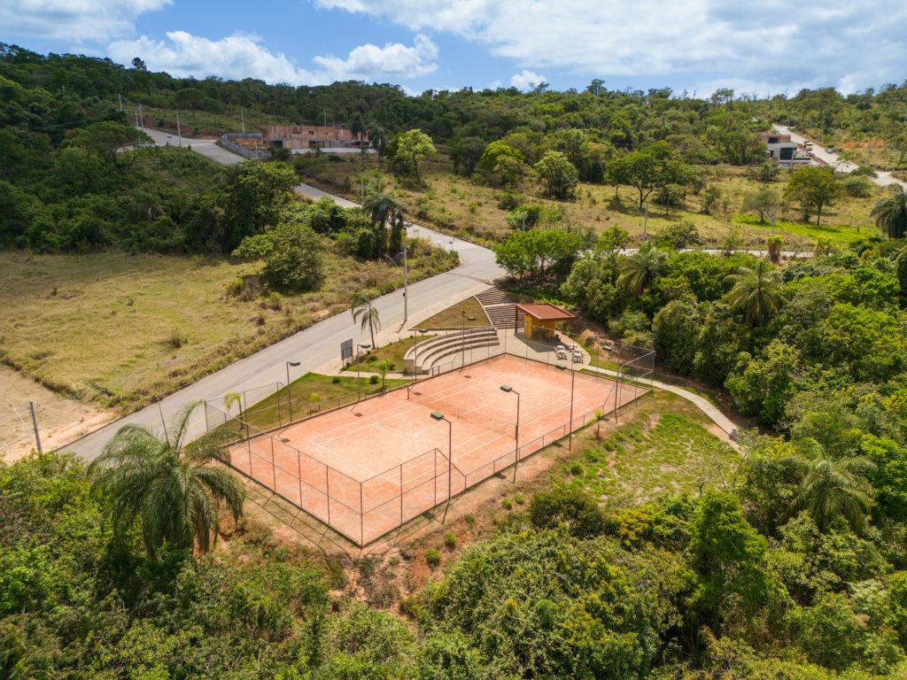 Foto de uma quadra de tenis de areia gradeada. Ao lado uma arquibancada e um quiosque de apoio com algumas mesas em área externa