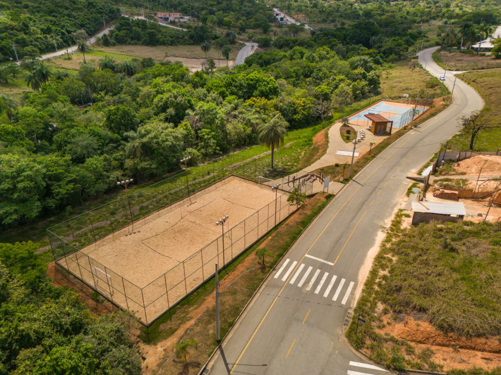 Foto de uma quadra de volei e futebol de areia,gradeada. Atrás um espaço com barras de alongamento e mais ao fundo quiosque de apoio com algumas mesas em área externa e quadra de futebol e basquete em concreto com pintura laranja e azul, gradeada.